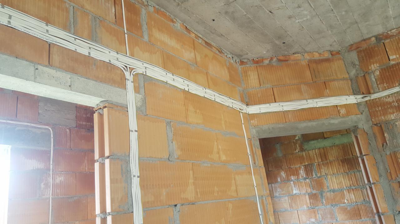 Surowe wnętrze budynku w trakcie budowy z widocznymi przewodami elektrycznymi ułożonymi wzdłuż ścian z cegły dziurawki.