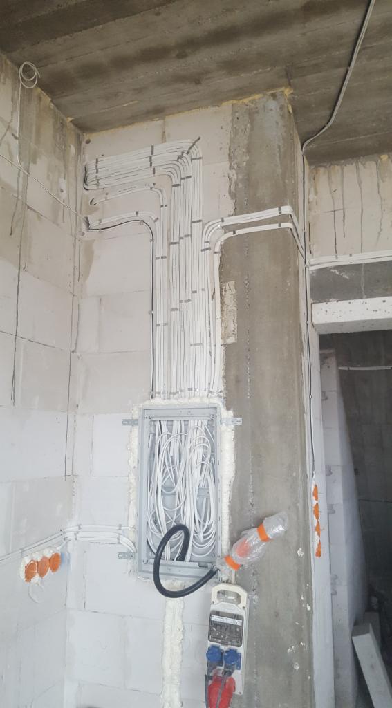 Rozprowadzenie przewodów elektrycznych w nowym budynku, kable w peszlach przymocowane do ściany, skrzynka rozdzielcza z dużą ilością przewodów, widoczne gniazdka i licznik elektryczny w folii...