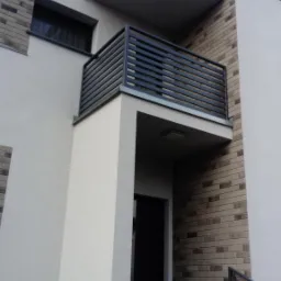 Nowoczesny balkon z ciemną, poziomą balustradą, widoczny z dołu, z fragmentem schodów wejściowych i elewacją budynku w tle.