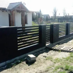 Nowoczesne ogrodzenie posesji z elementami z cegły klinkierowej i poziomymi przęsłami, widok z zewnątrz na tle domu jednorodzinnego w słoneczny dzień.