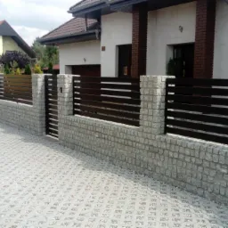 Nowoczesne ogrodzenie posesji z poziomych, ciemnych listew metalowych osadzonych na niskim murku z jasnej kostki granitowej, widok na dom z jasną elewacją i brązowymi elementami.