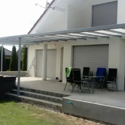 Szara metalowa pergola z przezroczystym zadaszeniem nad tarasem z betonowymi schodami, obok wanna SPA, w tle dom z szarymi roletami.