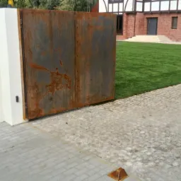Nowoczesna, rdzawa brama przesuwna z metalu w Śmigielu, zamontowana przy białym murze. W tle widoczny dom z czerwonej cegły i zielony trawnik.