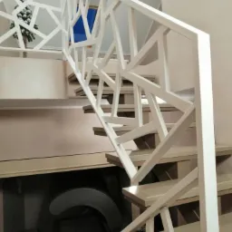 Nowoczesne schody z ażurową białą balustradą o geometrycznym wzorze, widok z dołu na górę, w tle mała choinka i niebieskie okno.