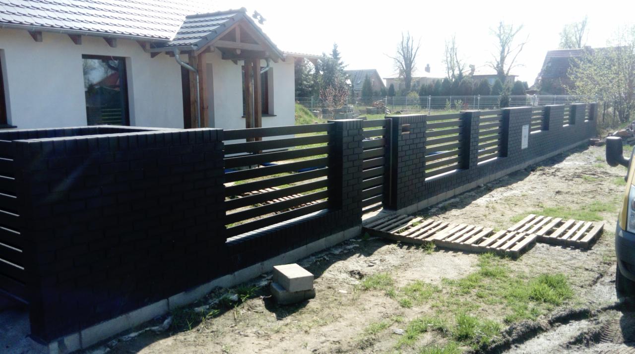 Nowoczesne ogrodzenie posesji z elementami z cegły klinkierowej i poziomymi przęsłami, widok z zewnątrz na tle domu jednorodzinnego w słoneczny dzień.