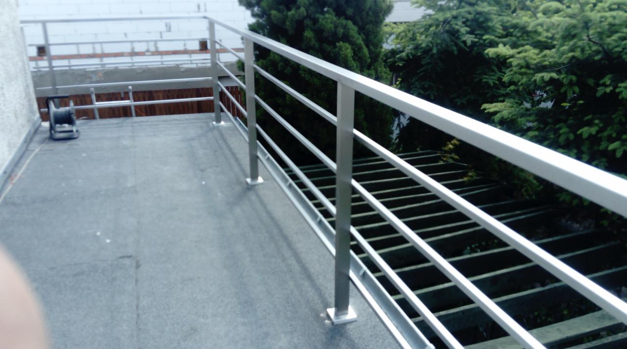 Barierka balkonowa ze stali nierdzewnej z widocznymi spawami u podstawy słupków, na tle zieleni i częściowo wykończonej ściany budynku.