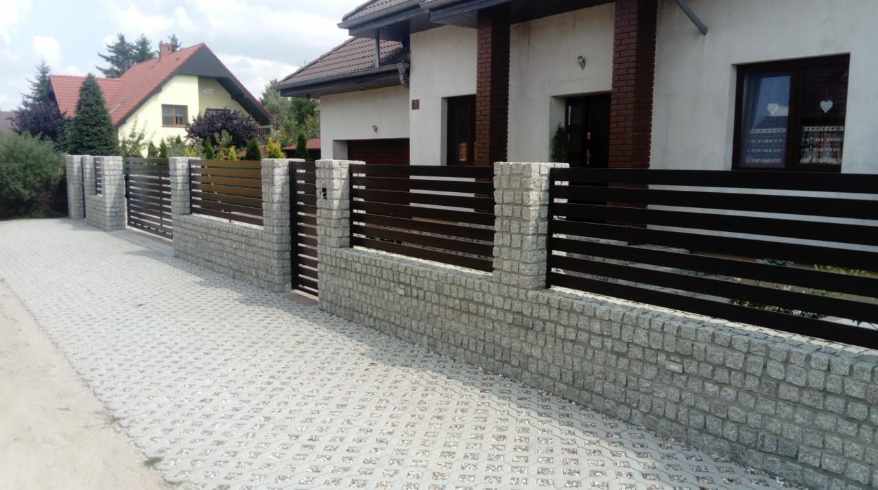 Nowoczesne ogrodzenie posesji z poziomych, ciemnych listew metalowych osadzonych na niskim murku z jasnej kostki granitowej, widok na dom z jasną elewacją i brązowymi elementami.