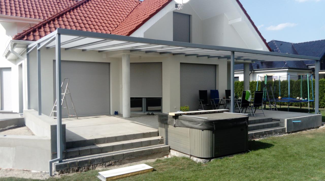 Szara pergola z zadaszeniem na betonowym tarasie przylegającym do domu z roletami zewnętrznymi, obok jacuzzi i schodów.