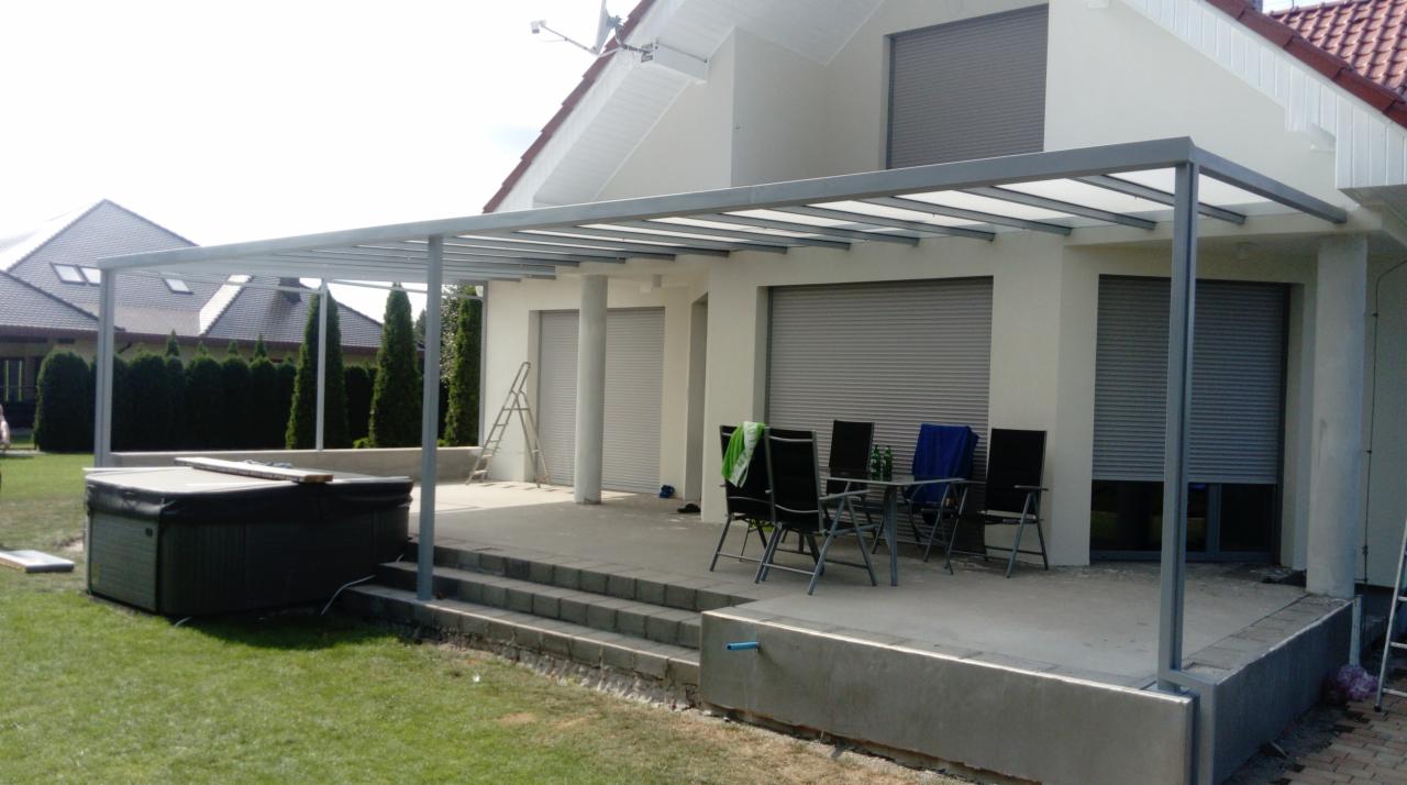 Szara metalowa pergola z przezroczystym zadaszeniem nad tarasem z betonowymi schodami, obok wanna SPA, w tle dom z szarymi roletami.