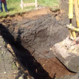 Wykop wąskiego rowu w ziemi wykonany koparką, widoczne warstwy gleby i fragment maszyny.