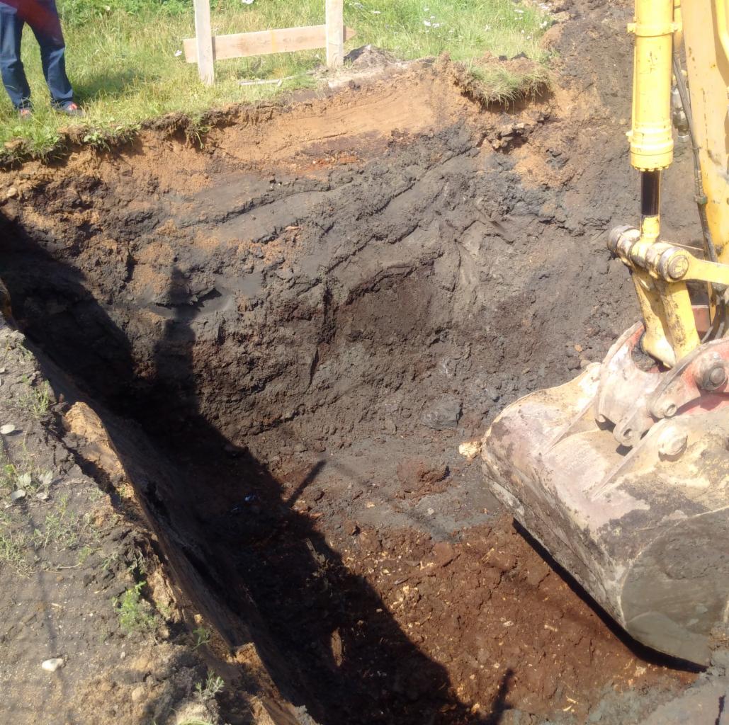 Wykop wąskiego rowu w ziemi wykonany koparką, widoczne warstwy gleby i fragment maszyny.