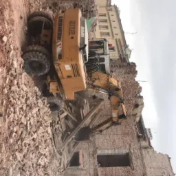 Żółta koparka Liebherr 902 w trakcie rozbiórki ceglanego budynku, z gruzem i fragmentami konstrukcji w tle, obok zaparkowana ciężarówka.