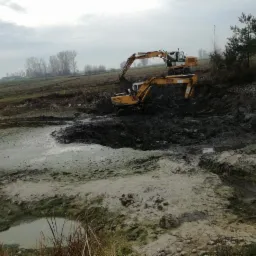 Dwie żółte koparki pracują w błotnistym wykopie, prawdopodobnie podczas pogłębiania lub tworzenia stawu w pochmurny dzień.