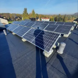 Panele słoneczne na płaskim dachu pokrytym papą, zamontowane na betonowych podstawach z widocznym cieniem osoby fotografującej, w tle zabudowa jednorodzinna i drzewa pod błękitnym niebem.