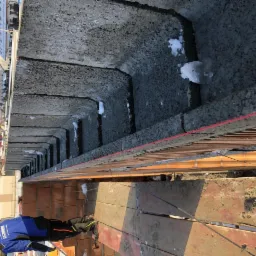 Wykonywanie ścian fundamentowych z bloczków betonowych, widoczne ślady zaprawy i niewielkie ilości śniegu w szczelinach, praca na wysokości zabezpieczona barierkami, pracownik w kombinezonie roboczym.