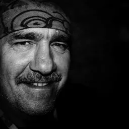 Portret uśmiechniętego mężczyzny z wąsami i bandaną na głowie, wykonany w czerni i bieli, z ciemnym tłem.