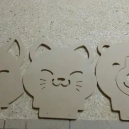 Trzy wycięte z jasnej płyty MDF kształty zwierząt (lis, kot, niedźwiedź) z wygrawerowanymi uśmiechniętymi pyszczkami, leżące na płycie wiórowej.