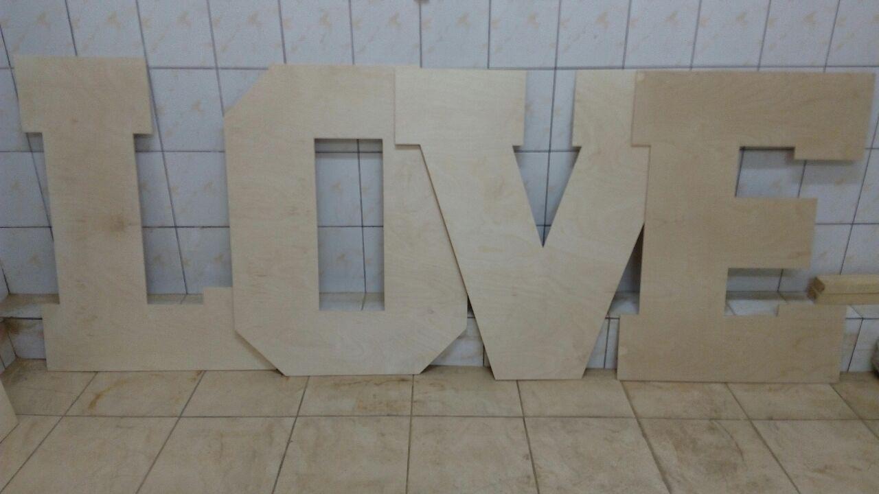 Surowe, drewniane litery LOVE, wycięte ze sklejki, ułożone na podłodze z płytek w warsztacie stolarskim.