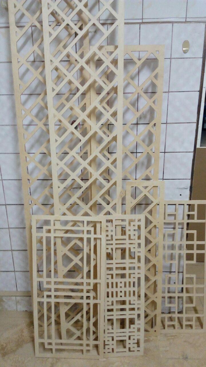 Surowe, drewniane panele dekoracyjne z wyciętymi wzorami geometrycznymi, o różnych rozmiarach i deseniach, ustawione pod ścianą z białych płytek.