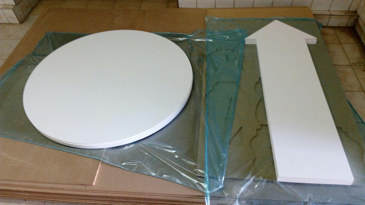 Biały okrągły blat i strzałka z drewna leżące na folii i kartonie, prawdopodobnie elementy dekoracyjne lub meblowe.