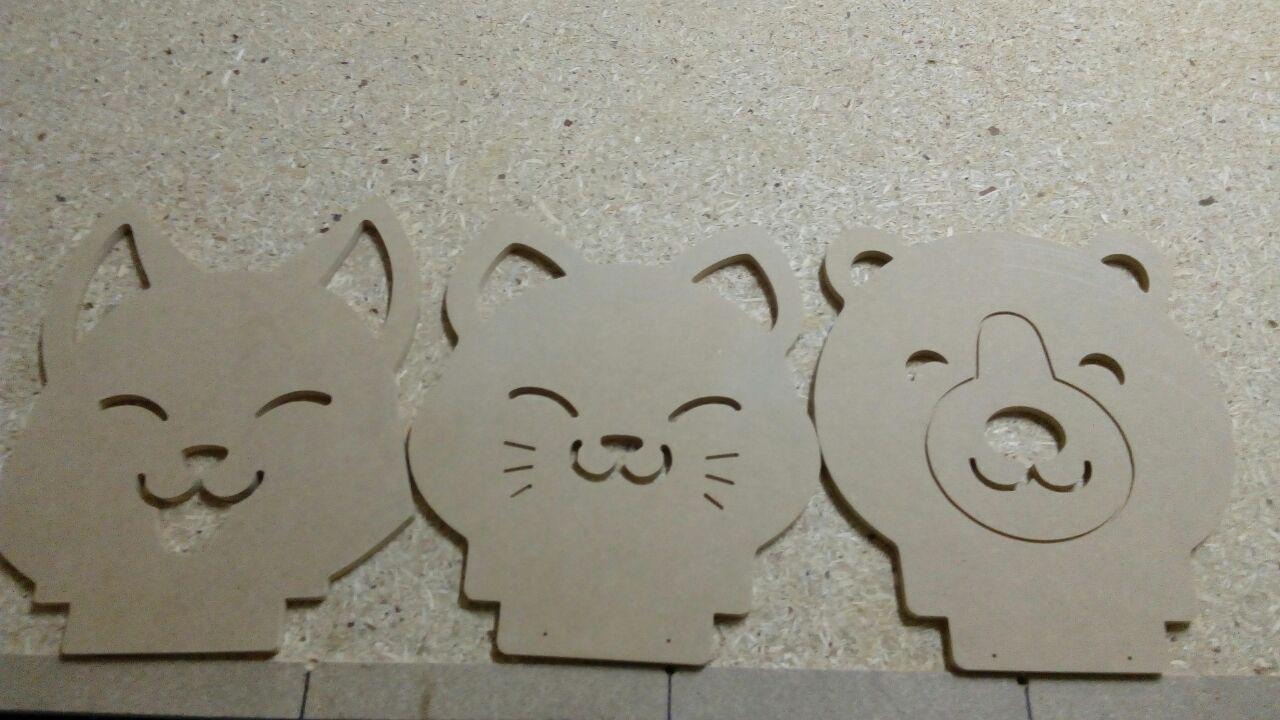 Trzy wycięte z jasnej płyty MDF kształty zwierząt (lis, kot, niedźwiedź) z wygrawerowanymi uśmiechniętymi pyszczkami, leżące na płycie wiórowej.