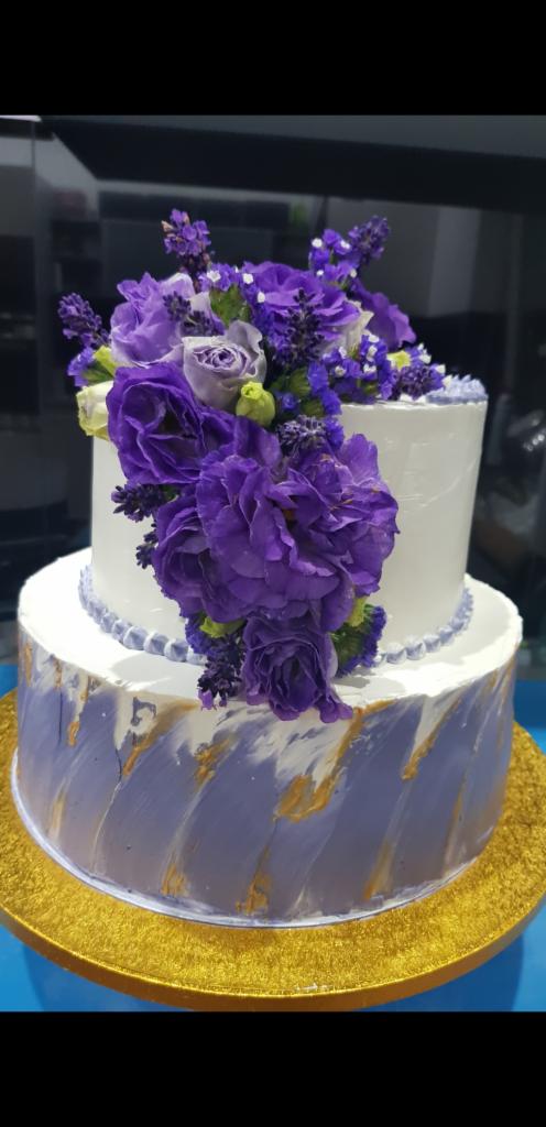 Dwupoziomowy tort z białym lukrem, zdobiony fioletowymi kwiatami i złotymi akcentami na dolnym poziomie, umieszczony na złotej tacy.