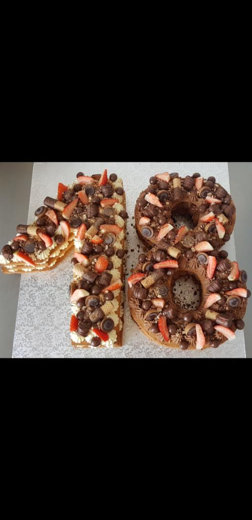 Tort w kształcie liczby 18, udekorowany kremem, kawałkami truskawek, czekoladkami i wafelkami, leżący na srebrnej podkładce.