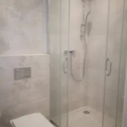 Nowoczesna łazienka z kabiną prysznicową z zaokrąglonymi szybami, wiszącą miską WC i płytkami w odcieniach beżu z delikatnym wzorem.