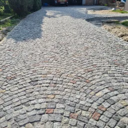 Ścieżka z kostki o nieregularnym kształcie ułożona w łukowy wzór, z elementami w odcieniach szarości i czerwieni, prowadząca do garażu w tle.