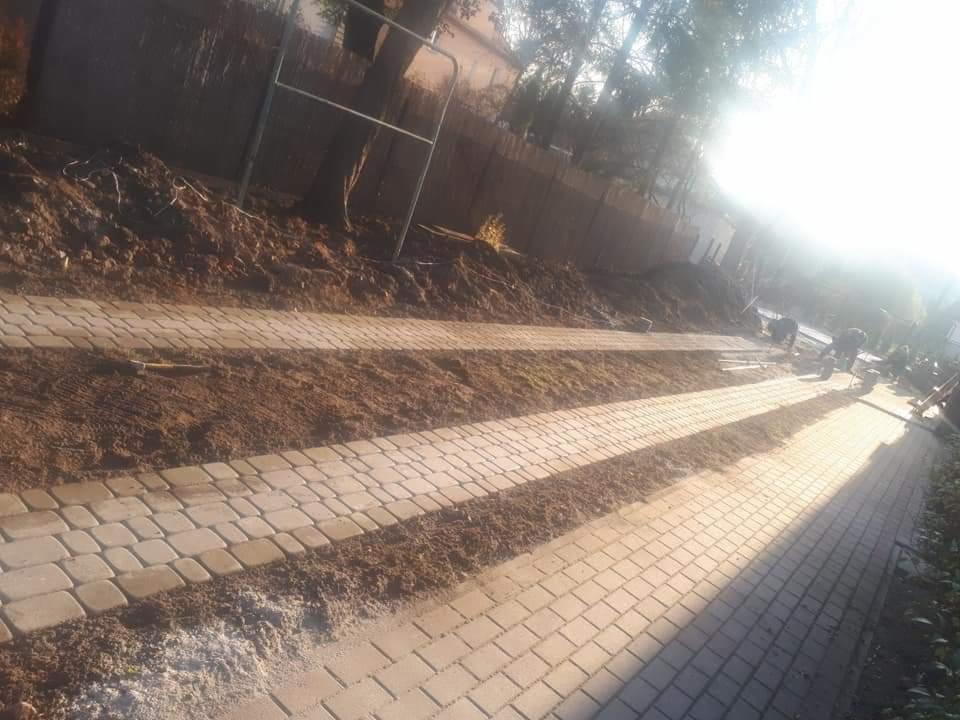 Układanie jasnej kostki brukowej wzdłuż posesji, widoczne niedokończone krawędzie i prace ziemne, silne światło słoneczne.