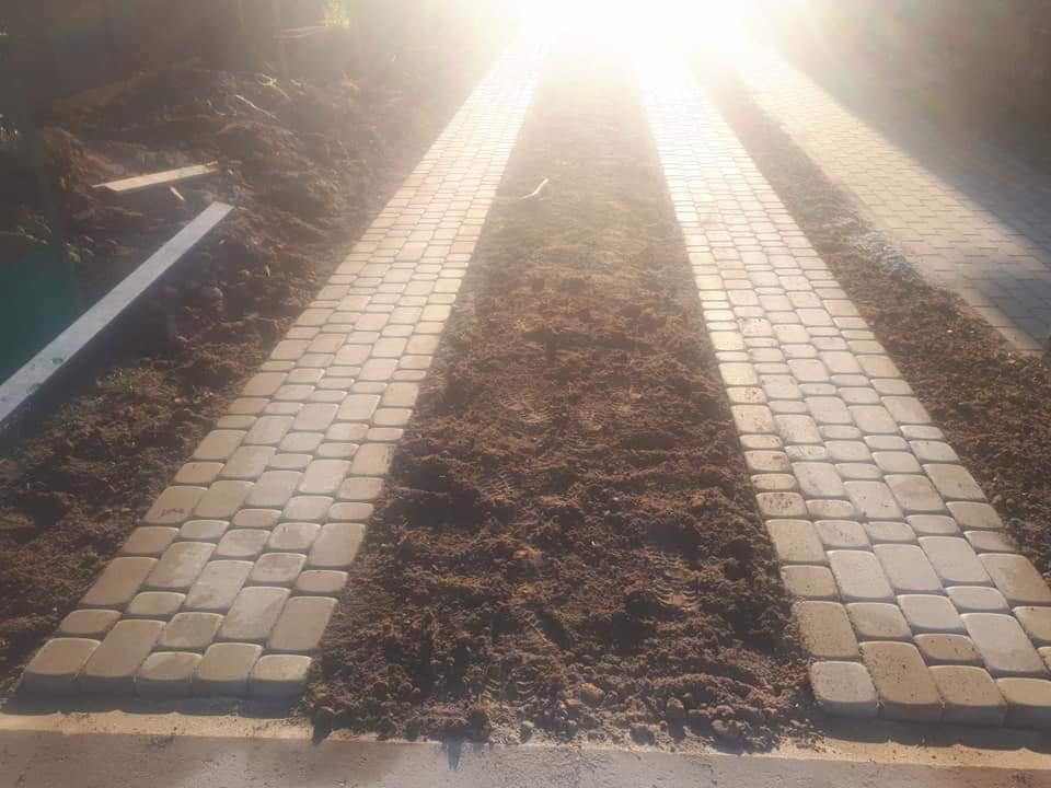 Świeżo ułożona ścieżka z jasnoszarej kostki brukowej, z widocznymi pozostałościami ziemi w spoinach i po bokach, w silnym oświetleniu słonecznym.