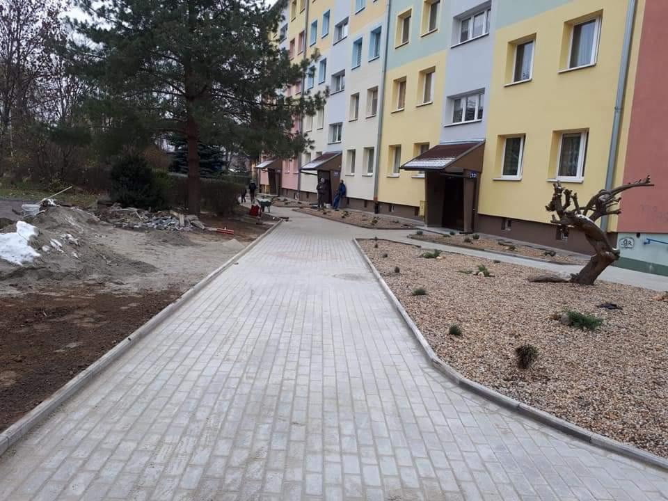 Nowy chodnik z jasnoszarej kostki brukowej przed blokiem mieszkalnym, z widocznymi pozostałościami po pracach ziemnych i dekoracyjnym nasypem z kamyków.