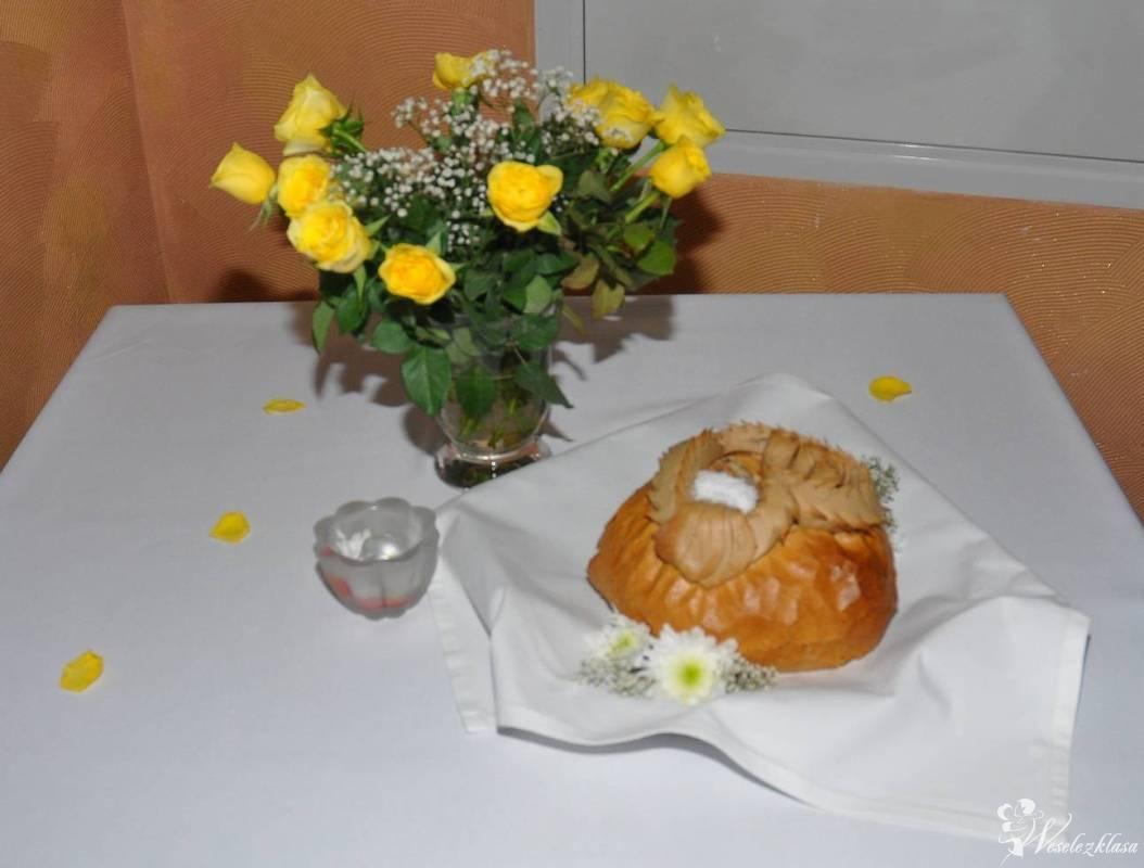 Uroczysty chleb weselny z solą na białym obrusie, obok bukiet żółtych róż w wazonie i dekoracyjny świecznik, całość dopełniają rozsypane płatki róż.
