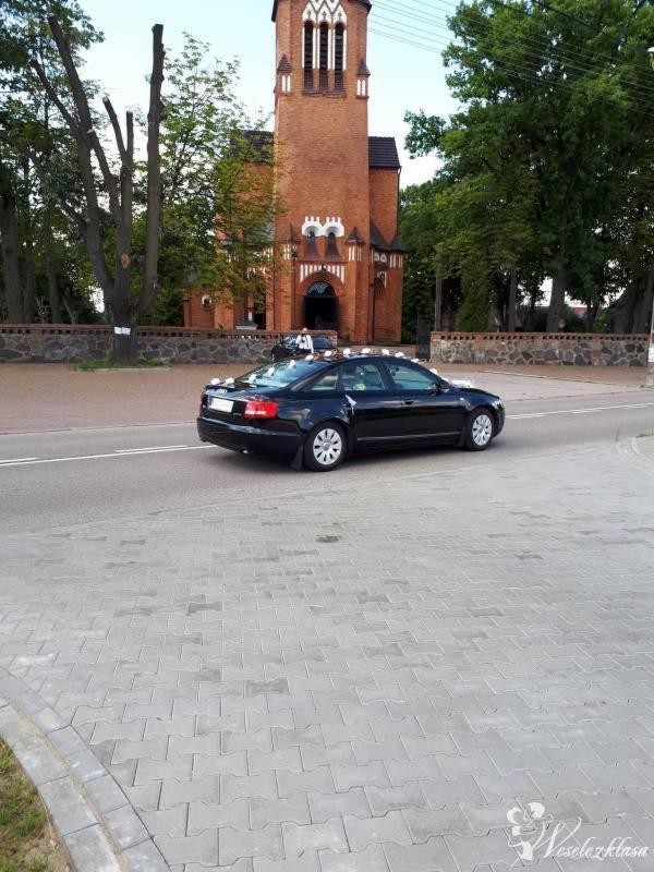 Czarny samochód udekorowany kwiatami przed ceglanym kościołem z wieżą, przygotowany do uroczystości ślubnej, widok z boku na tle drzew