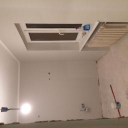 AT - Wnętrze w trakcie remontu: świeżo pomalowane ściany w jasnym odcieniu szarości, widoczne narzędzia malarskie przy ścianie, okno dachowe z grzejnikiem pod nim, ślady po wcześniejszych pracach...