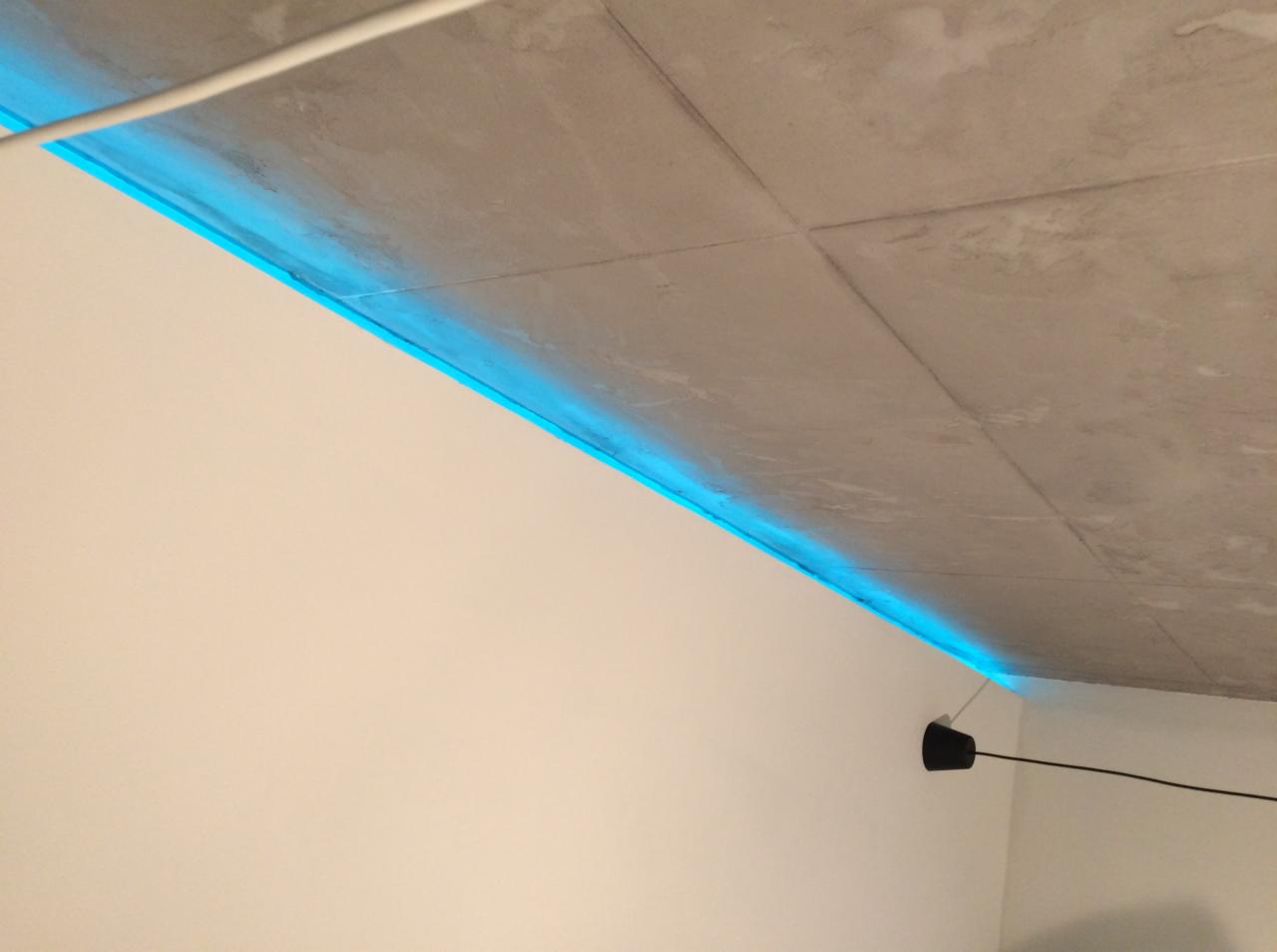 Betonowy sufit z podświetleniem LED w kolorze niebieskim, biała ściana i czarny element oświetleniowy.