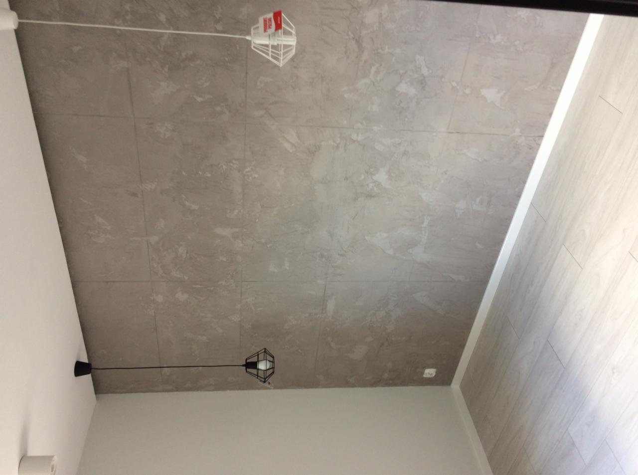 Szare płytki imitujące marmur na suficie, oświetlenie punktowe z czarnym abażurem i wisząca biała lampa druciana, jasne ściany i panele podłogowe.