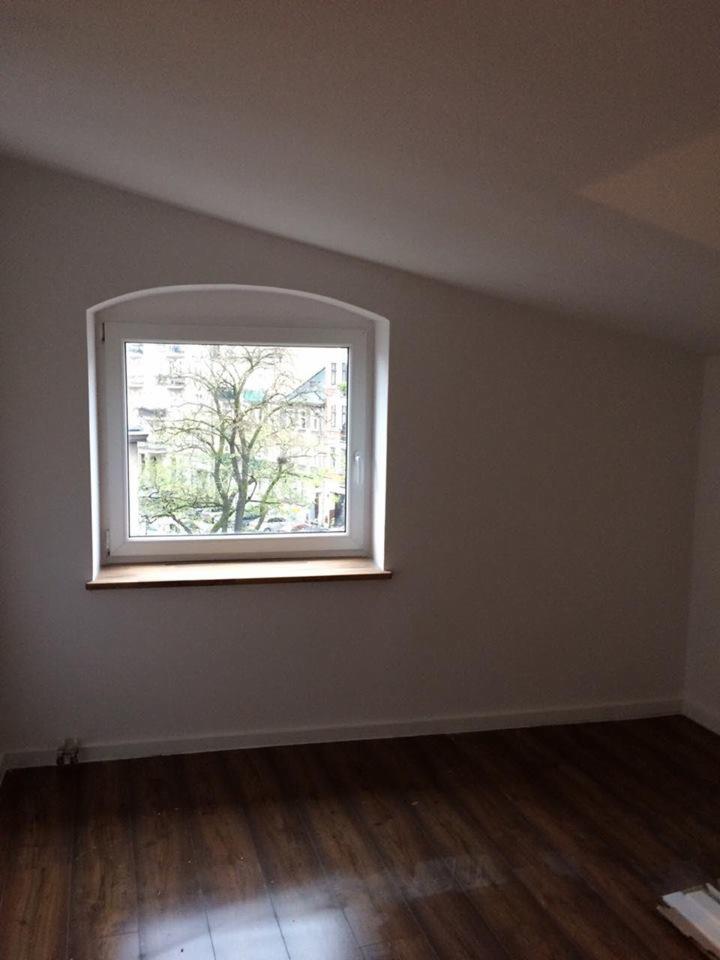 Pokój po remoncie z białymi ścianami i drewnianą podłogą, widok z okna na drzewo i budynki.