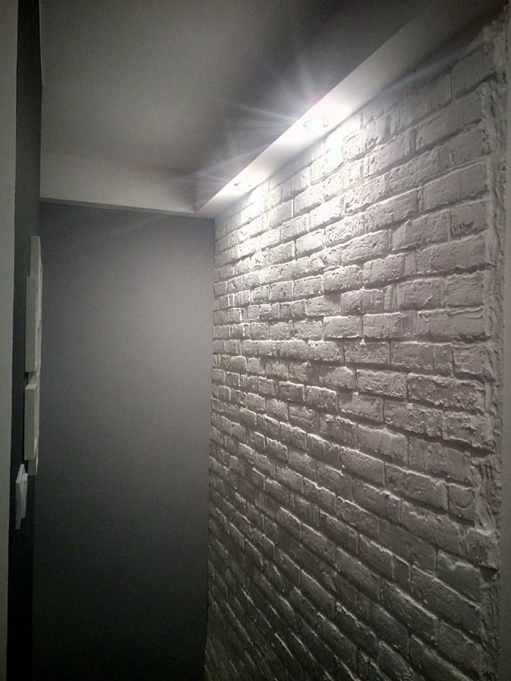 Wnętrze z dekoracyjną ścianą z cegły pomalowaną na biało, oświetloną punktowo, sąsiadującą z gładką szarą ścianą.