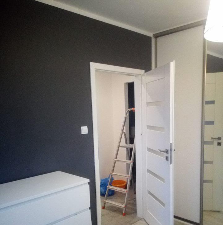 Ciemnoszara ściana po malowaniu, widoczna biała listwa przypodłogowa, drabina malarska z pomarańczowym wiadrem w przejściu do drugiego pomieszczenia, biała komoda i białe drzwi z przeszkleniami.
