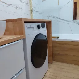 Nowoczesna łazienka w Oświęcimiu z zabudowaną pralką Beko, drewniany blat i obudowa, białe płytki imitujące marmur, szklany parawan nawannowy.