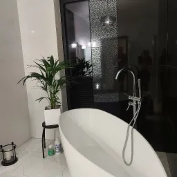 Nowoczesna łazienka z białą, wolnostojącą wanną, chromowaną armaturą, rośliną doniczkową na czarnym stoliku oraz czarno-białą mozaiką na ścianie.