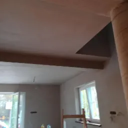 Wnętrze po nałożeniu gładzi gipsowej: sufit, ściany, kolumna owinięta taśmą, okna z widokiem na zewnątrz, drabina oparta o ścianę.