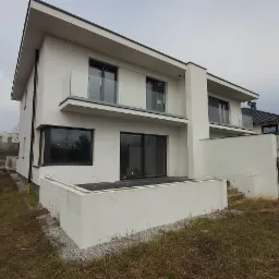 Nowoczesny, dwupiętrowy dom z białymi ścianami i szklanymi balustradami balkonów, widoczny z perspektywy ogrodu z niezagospodarowanym terenem.