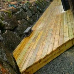 Drewniana kładka/podest prowadząca do wejścia, zbudowana na nierównym terenie, z widocznym kamiennym murem oporowym po lewej stronie. Deski ułożone diagonalnie, widoczne mocowania. Ujęcie z dołu...