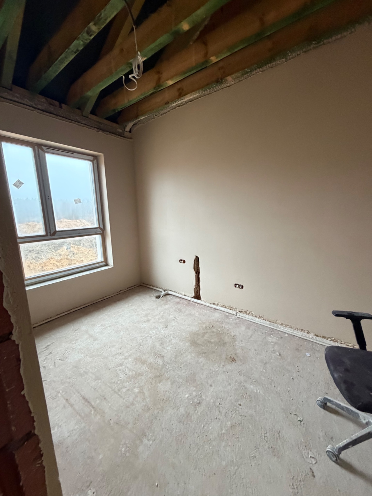 Pokój po tynkowaniu maszynowym: gładkie ściany, widoczne instalacje elektryczne, surowy stan podłogi i drewniany strop. Okno z widokiem na plac budowy.