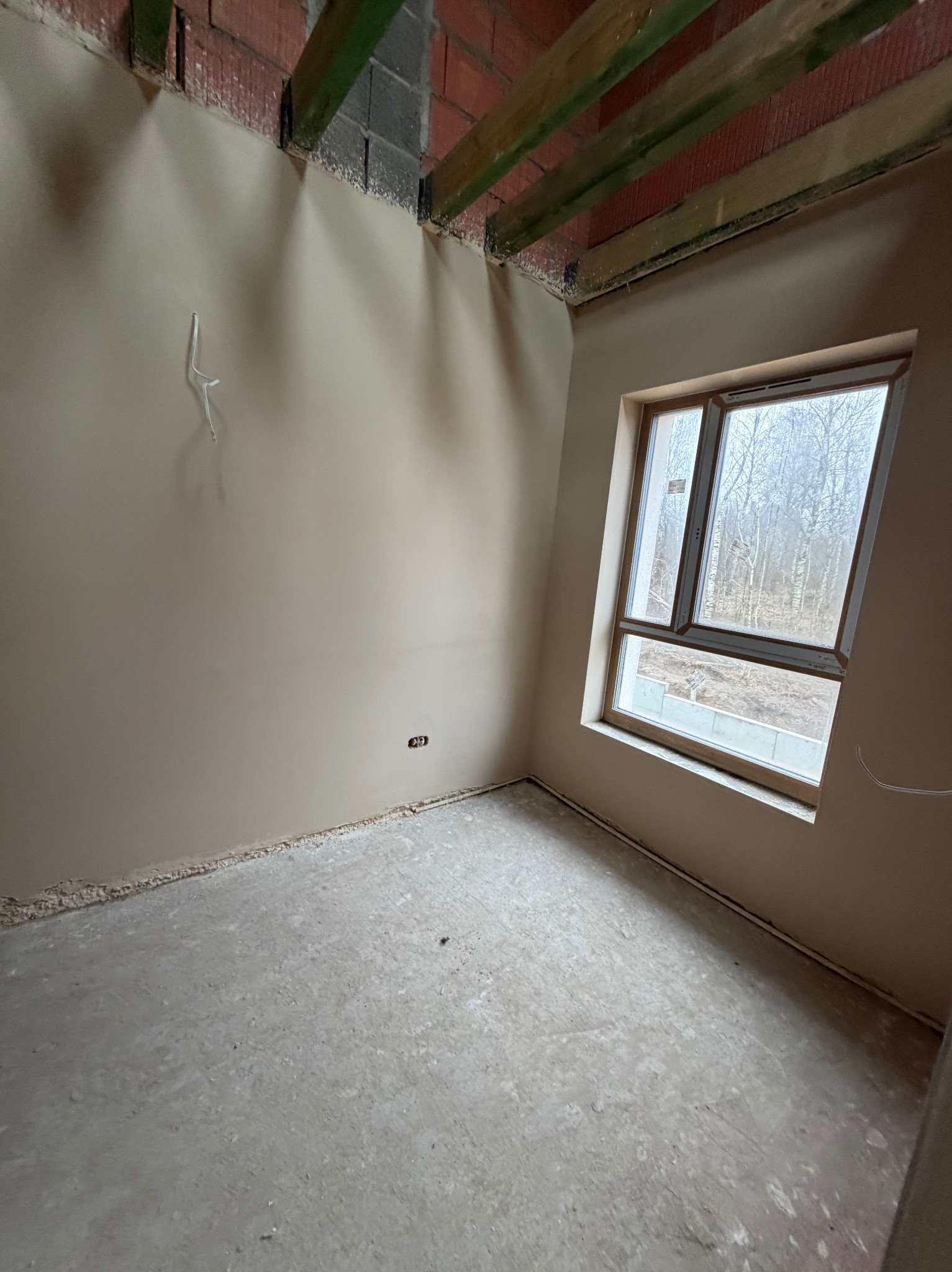 Wnętrze pokoju po nałożeniu gładzi, widoczne ściany w jasnym odcieniu beżu, okno z widokiem na drzewa i surowy beton na podłodze. Widoczne kable wystające ze ściany.