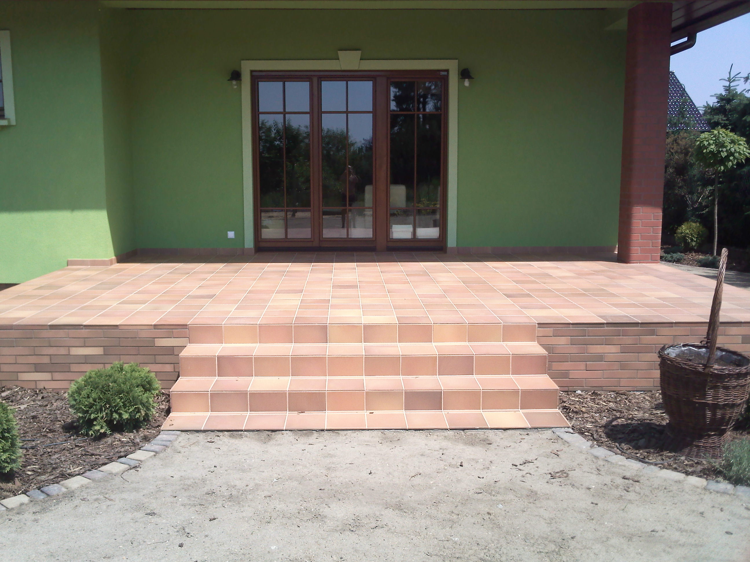 Wykończony taras z brązowych płytek ceramicznych z czterostopniowymi schodami, zielona ściana budynku w tle, widoczny kosz wiklinowy z ziemią.