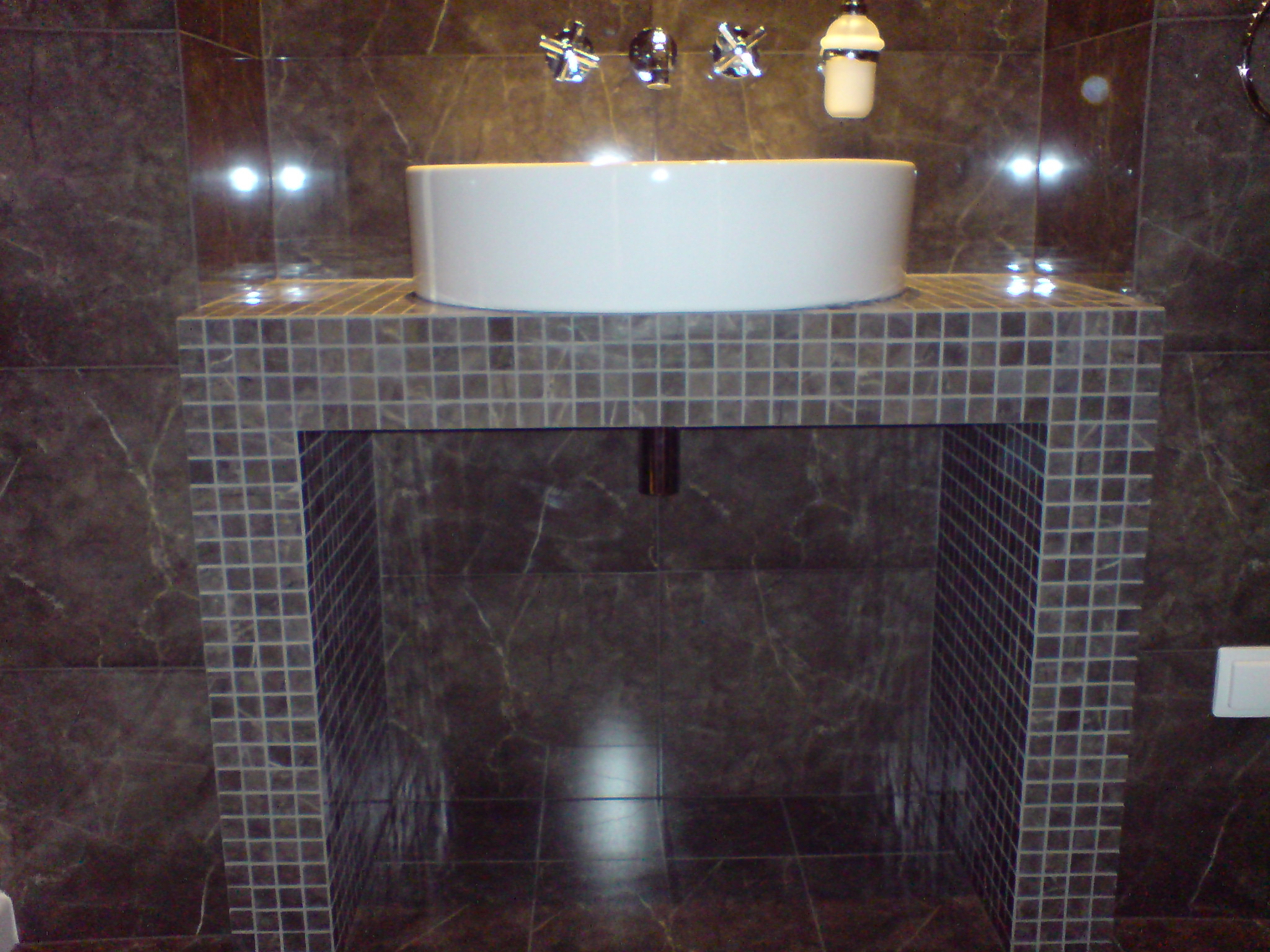 Umywalka nablatowa w łazience wykończonej ciemnymi, marmurkowymi płytkami i mozaiką. Bateria umywalkowa chromowana, trójotworowa.