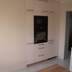 Jasna zabudowa kuchenna z piekarnikiem i mikrofalówką, czarne podłużne uchwyty, widoczny fragment drewnianego blatu na pierwszym planie.